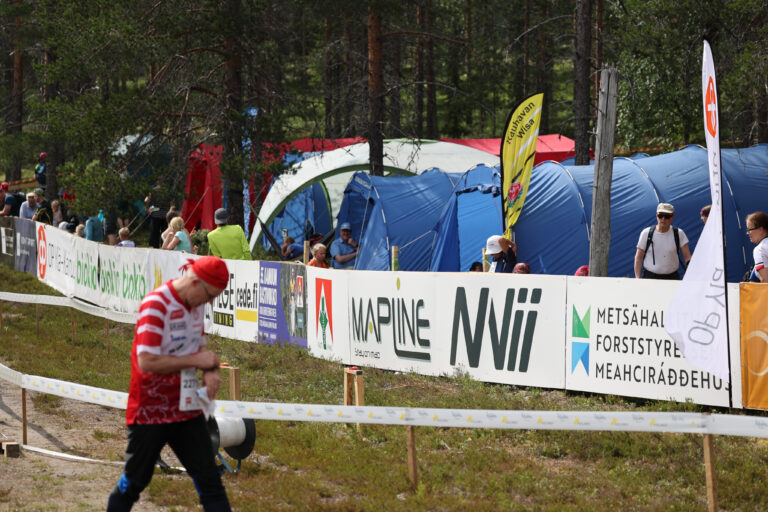 Punavalkoiseen asuun pukeutunut henkilö kävelee ulkoilmatapahtumassa suurten sinisten telttojen ja banderollien lähellä, samalla kun ihmiset seisovat ja istuvat telttojen lähellä metsäisellä alueella.