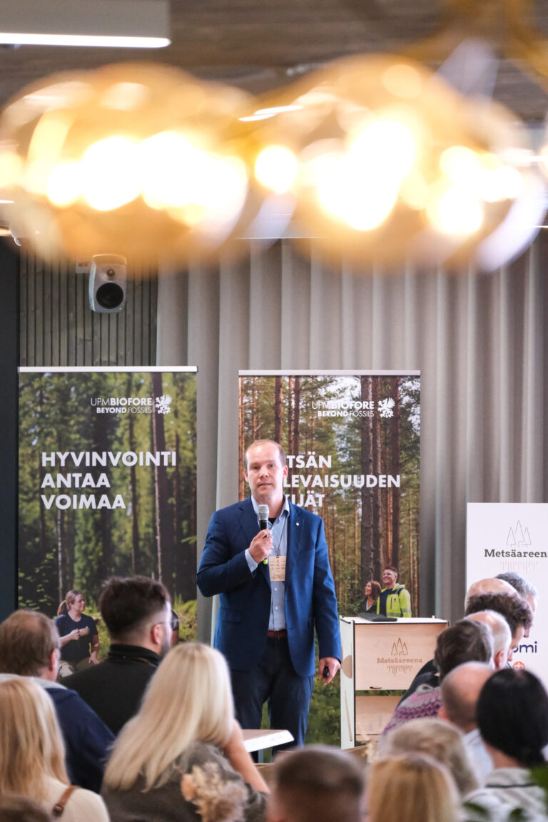 Siniseen pukuun pukeutunut mies puhuu mikrofoniin konferenssissa seisten bannereiden ja yleisön edessä. Lämmin valaistus ja sumea sisustus leijuvat täpötäynnä olevan huoneen yllä.