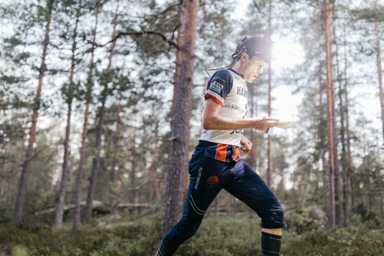 Urheiluvarusteisiin ja otsalamppuun pukeutunut henkilö juoksee metsän läpi kartta kädessään ja keskittyen suunnistukseen päivänvalossa. Korkeat puut ja vehreys ympäröivät urheilijaa.