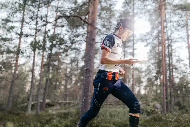 Urheiluvarusteisiin ja otsalamppuun pukeutunut henkilö juoksee metsän läpi kartta kädessään ja keskittyen suunnistukseen päivänvalossa. Korkeat puut ja vehreys ympäröivät urheilijaa.