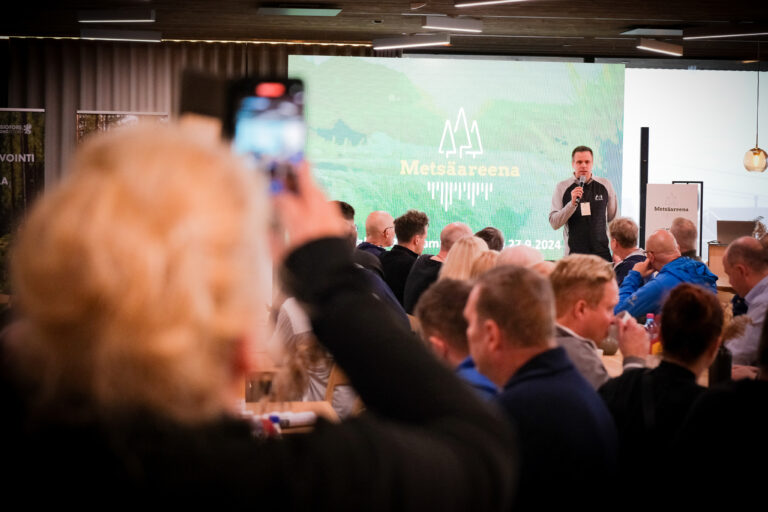 Mies puhuu lavalla Metsäareenan tapahtumassa istuvan yleisön kuunnellessa; etualalla oleva henkilö tallentaa puhujan älypuhelimellaan. Tapahtuman brändäys näkyy puhujan takana olevalla suurella näytöllä.