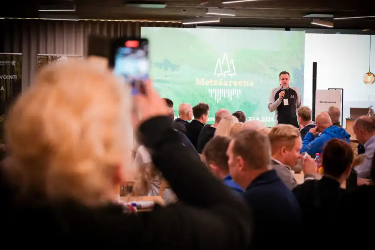 Mies puhuu lavalla Metsäareenan tapahtumassa istuvan yleisön kuunnellessa; etualalla oleva henkilö tallentaa puhujan älypuhelimellaan. Tapahtuman brändäys näkyy puhujan takana olevalla suurella näytöllä.