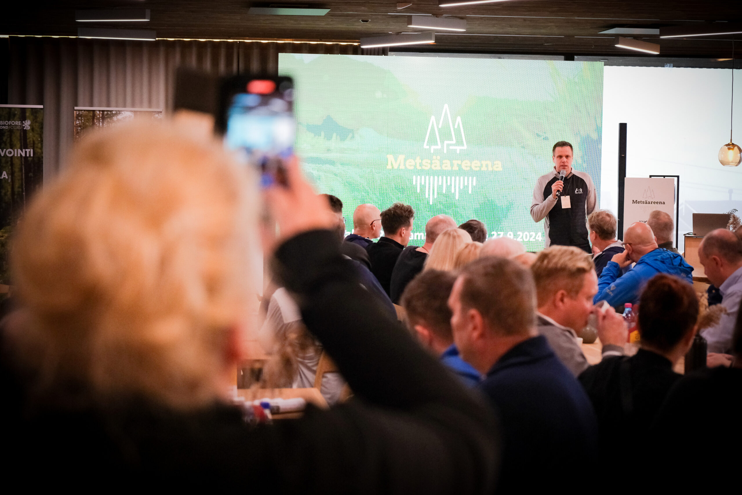 Mies puhuu lavalla Metsäareenan tapahtumassa istuvan yleisön kuunnellessa; etualalla oleva henkilö tallentaa puhujan älypuhelimellaan. Tapahtuman brändäys näkyy puhujan takana olevalla suurella näytöllä.