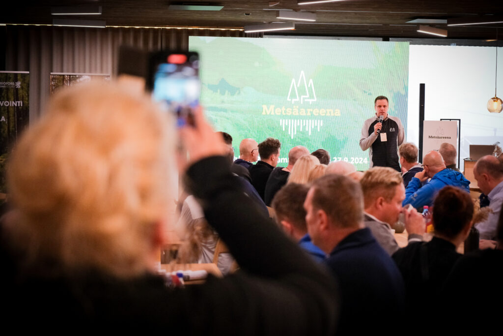 Mies puhuu mikrofoniin lavalla Metsäareenan tapahtumassa, hänen takanaan vihreä esitysnäyttö. Yleisö istuu ja kuuntelee, ja yksi henkilö etualalla pitelee puhelinta ottaakseen kuvan tai videon.