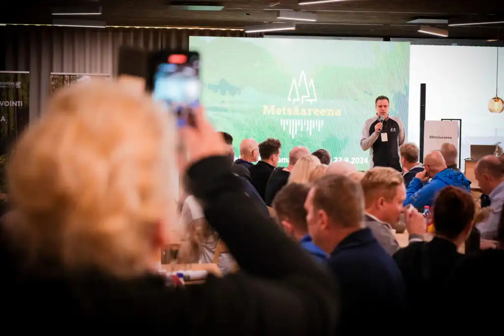 Mies puhuu mikrofoniin lavalla Metsäareenan tapahtumassa, hänen takanaan vihreä esitysnäyttö. Yleisö istuu ja kuuntelee, ja yksi henkilö etualalla pitelee puhelinta ottaakseen kuvan tai videon.