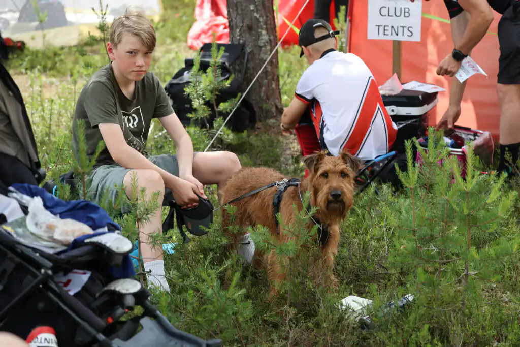 Vihreään paitaan ja shortseihin pukeutunut poika istuu nurmikolla ruskean koiran vieressä. Heidän takanaan toinen urheiluasuinen henkilö polvistuu punaisen teltan lähellä, jossa näkyy "CLUB TENTS" -kyltti. Heidän ympärillään on pensaita ja puita.
