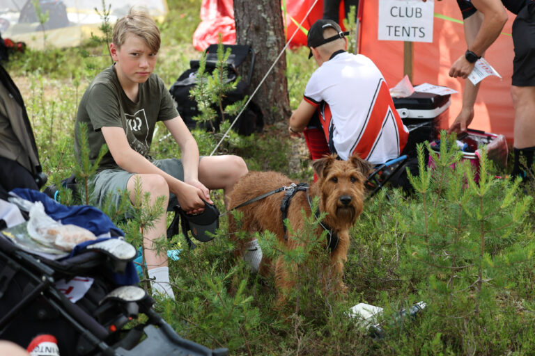 Vihreään paitaan ja shortseihin pukeutunut poika istuu nurmikolla ruskean koiran vieressä. Heidän takanaan toinen urheiluasuinen henkilö polvistuu punaisen teltan lähellä, jossa näkyy "CLUB TENTS" -kyltti. Heidän ympärillään on pensaita ja puita.