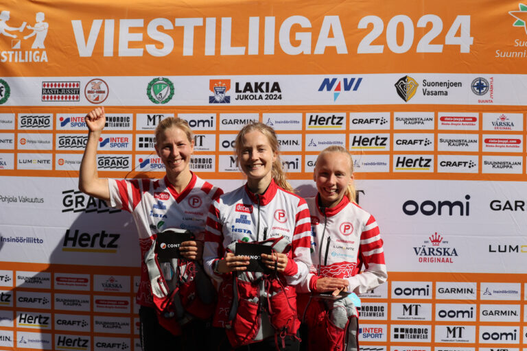 Kolme hymyilevää punavalkoisiin urheiluasuihin pukeutunutta naista seisoo tapahtuman taustan edessä, jossa lukee "Viestiliiga 2024", pitelevät pokaaleja ja poseeraavat ylpeänä valokuvaa varten.