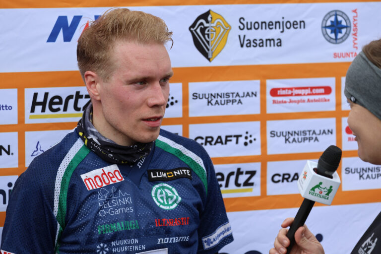 Siniseen urheilupaitaan pukeutunutta miesurheilijaa haastattelee mikrofonia pitelevä toimittaja. Tapahtuman sponsorien logot näkyvät taustalla olevassa bannerissa.