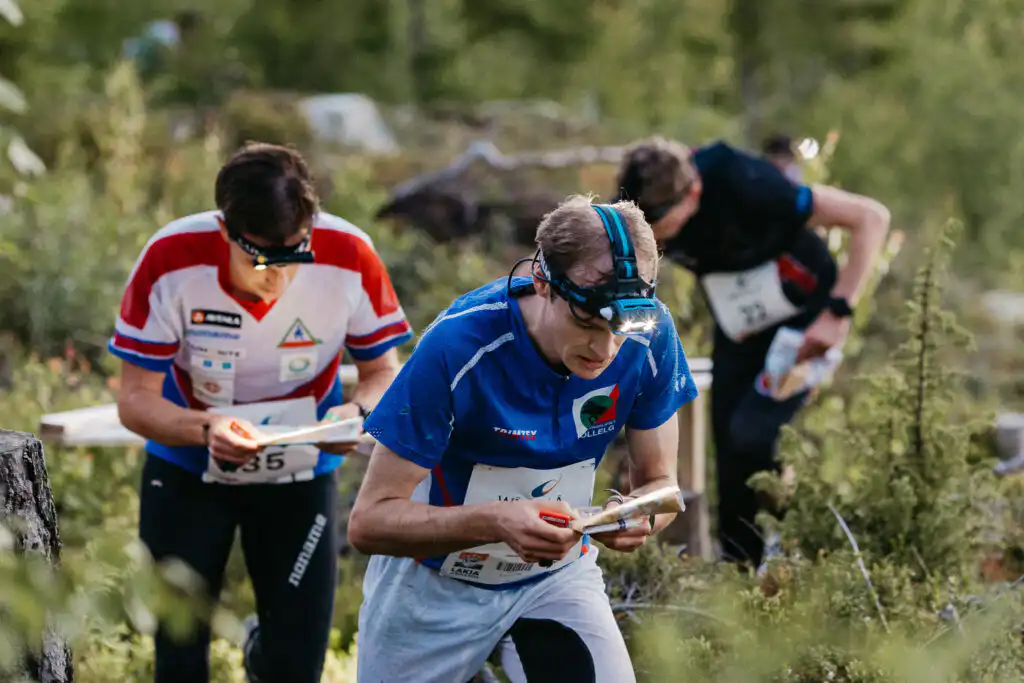 Kolme ulkoiluvaatteissa ja otsalampuissa olevaa urheilijaa keskittyy suunnistamaan karttojen avulla suunnistuskilpailussa metsäalueella. Vehreä kasvillisuus ympäröi heitä juostessaan.