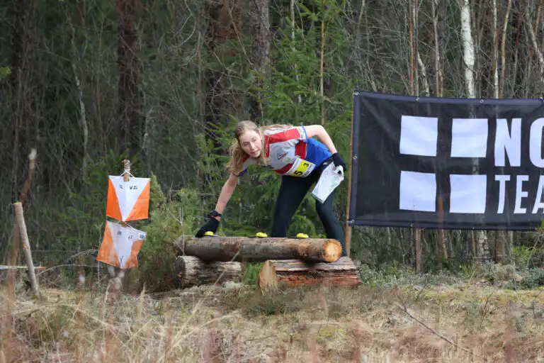 Urheiluvaatteissa oleva nainen kumartuu tukin yli ja tarkistaa suunnistuslippua metsäalueella. Hänellä on kartta ja kilpailunumerolappu mustan, valkoisella tekstillä ja neliöillä varustetun kyltin vieressä.