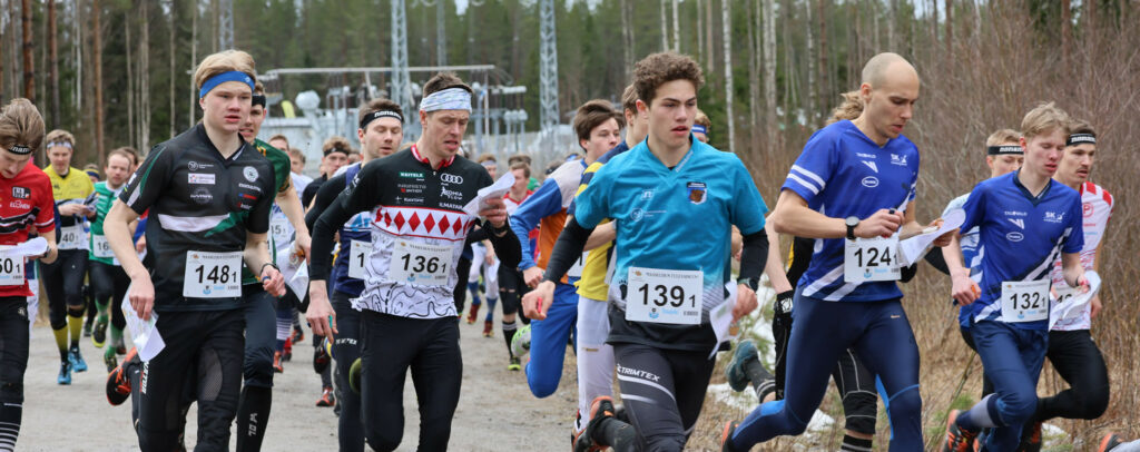 Ryhmä juoksijoita, enimmäkseen nuoria miehiä, aloittaa kilpailun ulkona puiden ympäröimällä hiekkapolulla. Heillä on numeroidut numerolaput ja urheiluvaatteet, ja joillakin on suunnistuskartat. Tausta on hieman sumea.