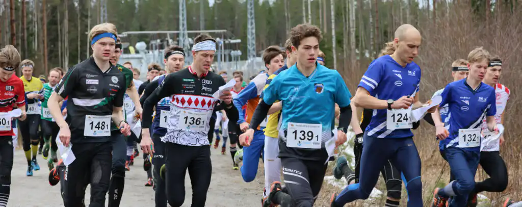 Ryhmä juoksijoita, enimmäkseen nuoria miehiä, aloittaa kilpailun ulkona puiden ympäröimällä hiekkapolulla. Heillä on numeroidut numerolaput ja urheiluvaatteet, ja joillakin on suunnistuskartat. Tausta on hieman sumea.
