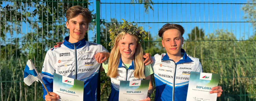 Kolme nuorta urheilijaa samanlaisiin sinivalkoisiin urheiluasuihin pukeutuneena hymyilevät ja pitelevät diplomejaan. Keskellä olevalla tytöllä on kukkakruunu. Vasemmalla on Suomen lippu, ja taustalla näkyvät vihreä aita ja puita.