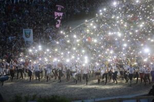 Suuri ihmisjoukko juoksee kohti kameraa kilpaa, ja heidän takanaan on paljon katsojia. Lukuisat kirkkaat valot, jotka ovat todennäköisesti peräisin kameran salamavaloista tai puhelimen taskulampuista, valaisevat kohtausta.
