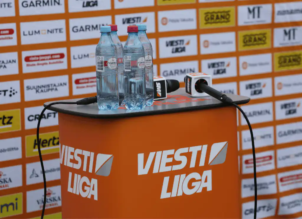 Oranssilla korokkeella, jossa on Viesti Liiga -merkki, on mikrofoneja ja kaksi vesipulloa, ja taustalla on erilaisia sponsorien logoja.