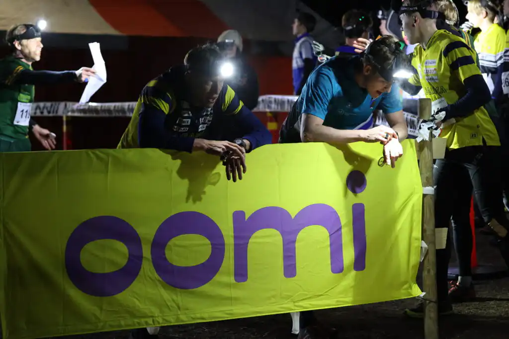 Osallistujat otsalampuissa nojaavat kirkkaankeltaisen banderollin yli, jossa on violetilla "oomi", ja vaikuttavat keskittyneiltä ja sitoutuneilta, kun muut lukevat lehtiä lähellä yöllisessä ulkoilmatapahtumassa.
